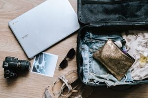 Flat lay of travel essentials including a laptop, a camera, sandals, sunglasses, a polaroid, and a bag full of clothes and personal accessories | Luxury Homes by Brittany Corporation