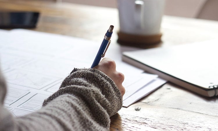 College student writing and answering homework on a wooden table with a mug of coffee | Luxury Homes by Brittany Corporation