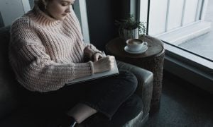 A young woman in a beige knitted sweater writing down her thoughts in her journal | Luxury Homes by Brittany Corporation