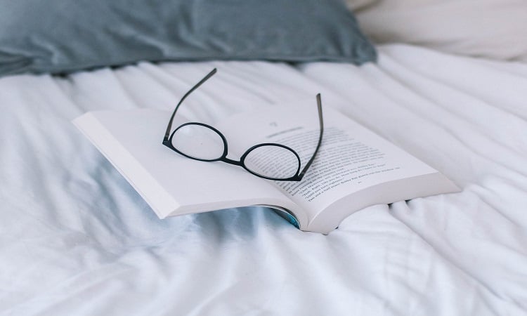 A pair of reading glasses resting on top of a book on a bed with white sheets and a grey pillow in a luxury house | Luxury Homes by Brittany Corporation