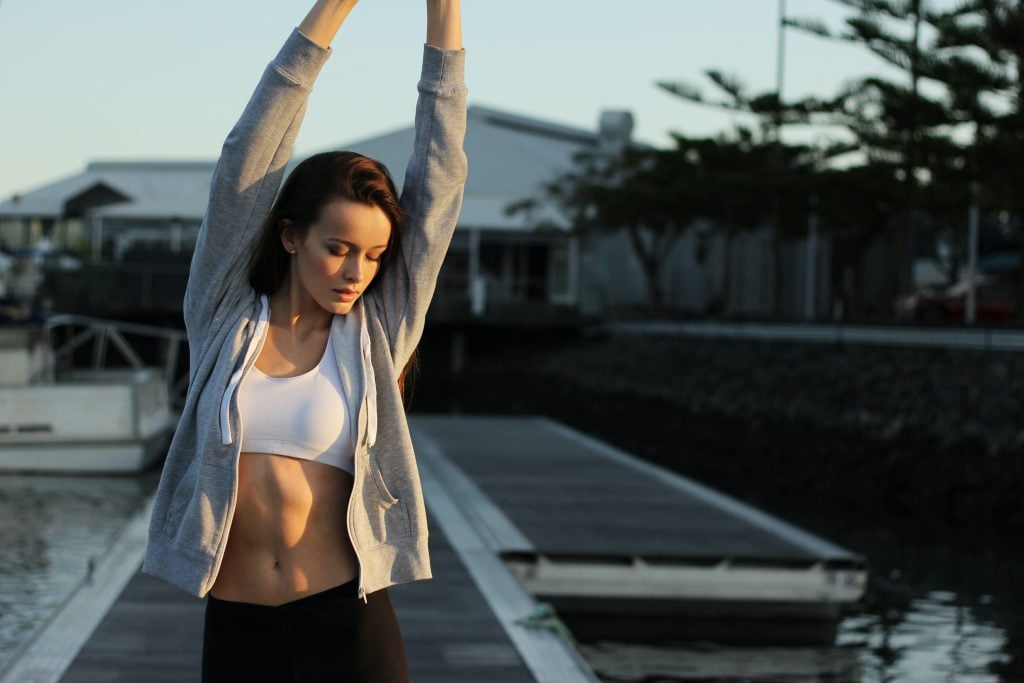 Young Caucasian woman in white sports bra, grey jacket, and black pants stretching her arms after her morning workout in her luxury themed community | Luxury Homes by Brittany Corporation