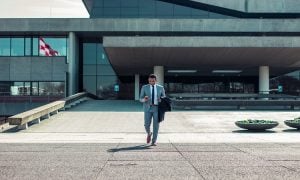 Young caucasian professional with a mixed ethnic background in a grey suit using his phone while stepping out of his office building where he works for a real estate developer | Luxury Homes by Brittany