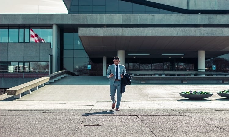 Young caucasian professional with a mixed ethnic background in a grey suit using his phone while stepping out of his office building | Luxury Homes by Brittany