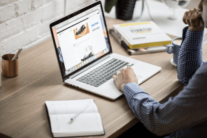 A man in his luxury home wearing a blue checkered polo shirt browsing the internet on his silver laptop which is placed on a brown wooden table alongside his notebook with a pen