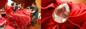 A picture divided into 2 separate images; on the left, a silk chiffon scarlet red draped dress with four asian malaysian workers weaving or sewing small diamonds into the dress on a long table inside a spacious brown studio room; on the right is a zoom in image of a pear cut or pear shape sparkling diamond sewed into the dress and reflecting the light in every edge