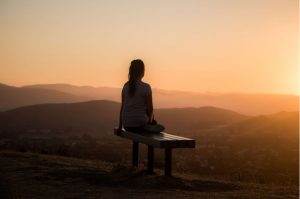 Woman practicing self-care by sitting on a bench and watching the sunset view above mountaintop scenery- Luxury homes by Brittany