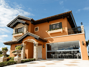 Orange two floor house with Italian architecture, marble flower pots, bricked pillars, and a glass wall showing the interiors of a family dining area | Luxury Homes by Brittany Corporation