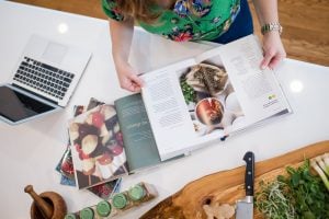 Top view of light skinned brunette woman wearing tropical colors, reading through books on plant-based diets on a white counter with healthy ingredients and a chopping board and a macbook laptop, in a kitchen | Luxury Homes by Brittany Corporation