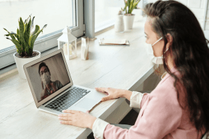 Caucasian brunette female in her twenties wearing a pink button down shirt and a face mask, video calling an Asian male wearing a face mask, through her macbook laptop, on a windowside wooden desk with pots of succulent plants and home decoration | Luxury Homes by Brittany Corporation