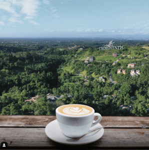 A cup of coffee on a saucer with a teaspoon, on a wooden table, overlooking a landscape of a pine tree forest, dotted with mansions, and a huge “Crosswinds” sign, perfect vacation property | Luxury Homes by Brittany Corporation