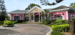 Pink Southern American style club house by a paved rotunda with healthy green pine trees, palm trees, and bushes | Luxury Homes by Brittany Corporation