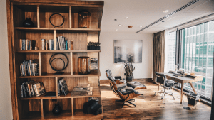 Spacious office on a high floor in a building, with a wooden corner shelf of books and ornaments, a leather recliner armchair, a wall painting, a potted green snake plant, a leather computer chair together with a wooden desk against the glass wall overlooking a view from high up | Luxury Homes by Brittany Corporation