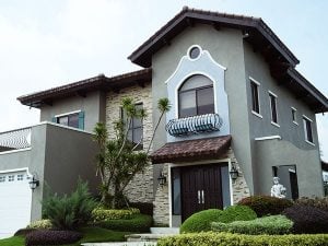 Grey two floor mansion with Italian architectural details and warm colored bricks, stairs leading to the elegant doorway, and a terrace on top of an indoor carport garage, surrounded by a healthy green garden of bushes, flowers, palm trees, and a marble statue | Luxury Homes by Brittany Corporation