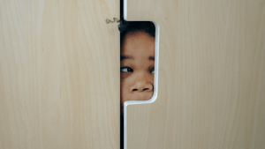 Dark skinned young little girl with curly black hair hiding in a light colored wooden cabinet | Luxury Homes by Brittany Corporation