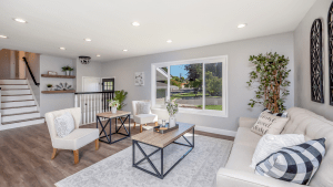 Well lit living room in neutral colors and wooden flooring, with wooden coffee tables, comfortable white couches with textured pillows, on a thick soft carpet, and a small leafy indoor tree plant in the corner, as well as a window overlooking the neighborhood | Luxury Homes by Brittany Corporation