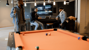 Three black men in hip and casual attire drinking whiskey in an indoor bar and playing billiards on an orange pool table, while music is playing and flashed on a flat screen TV installed on the wooden wall, with two speakers on both sides | Luxury Homes by Brittany Corporation