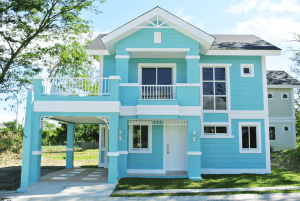 Blue western picket fence house with a terrace on top of an open carport garage, in a green grassy lot surrounded by trees | Luxury Homes by Brittany Corporation