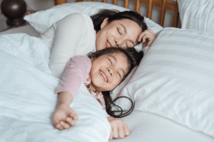 Beautiful Asian mother cuddling with her happy daughter comfortably in a white bed, under a thick duvet or comforter | Luxury Homes by Brittany Corporation