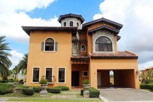 Orange Italian ready-for-occupancy mansion with a brick tower; a front garden with palm trees, bushes, and potted ferns; an open garage or carport; under a cloudy blue sky | Luxury Homes by Brittany Corporation