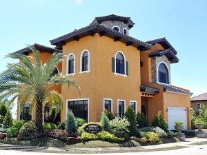 Italian Orange mansion with a brick tower; a front garden with palm trees, bushes, and potted ferns; an gated garage or carport; under a clear blue sky | Luxury Homes by Brittany Corporation