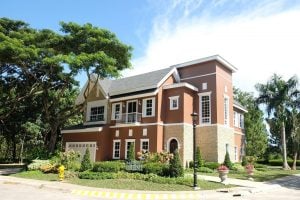 Red bricked two floor mansion with high ceiling in Old English fashion with a white picket fence balcony and an indoor carport garage, and a healthy grassy mini garden with pink flowers, on a corner lot surrounded by palm trees and other trees | Luxury Homes by Brittany Corporation