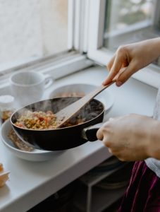 Fair skinned hands placing vegetables in a white bowl from a black pan with a wooden spatula, showing a plant-based diet on the white table | Luxury Homes by Brittany Corporation 