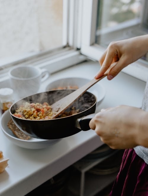 Fair skinned hands placing vegetables in a white bowl from a black pan with a wooden spatula, showing a plant-based diet on the white table | Luxury Homes by Brittany Corporation