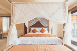 White queen size bed with orange pillows and a wooden bed frame with thin white drapes in a spacious bedroom with an orange couch and a curtained doorway leading to an outdoor courtyard  | Luxury Homes by Brittany Corporation