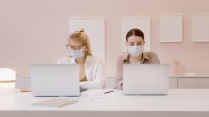 Two women working on their laptops while socially distanced and with masks on as an effect of the Covid-19 pandemic | Luxury Homes by Brittany Corporation