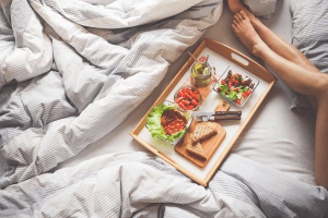 Olive skin tone legs next to a wooden tray with a healthy green breakfast of salads, fruits and berries, whole wheat sandwiches, and a mason jar of green tea with a metal straw, on a king size bed with striped blue and white duvet | Luxury Homes by Brittany Corporation