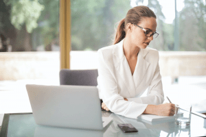  Beautiful brunette woman with glasses and red lipstick, wearing a white blazer while jotting down notes during an online webcam meeting through her laptop | Luxury Homes by Brittany Corporation