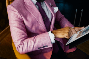 Formal attire of thick pink tweed coat over a polka dotted shirt and violet tie on a tan man with a classy watch and accessories, using his iPad for browsing the internet | Luxury Homes by Brittany Corporation