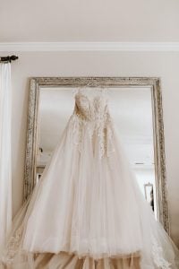 Dainty peachy white classic wedding dress in layers of tulle and embroidered classic details, hanging from a clear plastic hanger hooked on to a tall grand mirror with a classic frame inside an elegant room