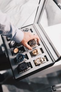 Hand of a man wearing a long sleeved formal shirt, reaching for a classic expensive watch from a leather case collection of other branded timepieces like Rolex | Luxury Homes by Brittany Corporation