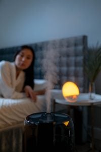 A black humidifier puffing out steam inside a bedroom in the evening, with a background of a woman wearing a robe in bed, next to a night lamp on a beside table