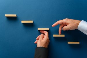 Hand of a businessman supporting wooden steps for his partner to walk his fingers upwards in a conceptual image of business partnership. Over blue background. Tips to succeed in real estate | Luxury Homes by Brittany Corporation