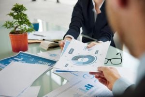 Man and woman exchanging company or financial reports over a meeting, wearing professional attire, on a glass desk with a bonzai tree plant, in a white office | Luxury Homes by Brittany Corporation