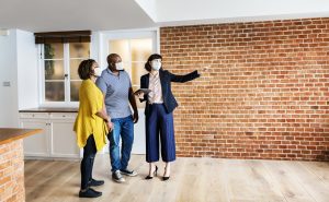 A black couple being toured by a white real estate agent in a professional outfit, around a modern industrial home with bricked walls and white kitchen | Luxury Homes by Brittany Corporation