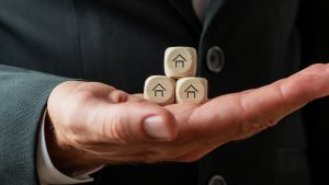 Hand of a businessman or realtor holding three wooden dices with house shapes on them | Luxury Homes by Brittany Corporation