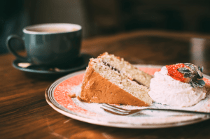 A plate of sliced strawberry shortcake with strawberry on icing, next to a cup of hot coffee, all on a wooden table - Luxury homes for sale - Brittany Corporation