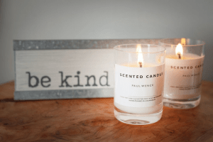 Branded scented lit candles with simple packaging, next to a sign that says be kind