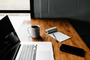 Coffee mug on a wooden table in the living room of a luxury home which is featuring a notepad with a pen a phone and a silver Apple laptop | Luxury Homes by Brittany Corporation