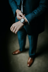 Young professional male adjusting in a navy blue three piece suit with brown leather shoes adjusting his watch before getting ready to leave his luxury home in the Phlippines | Brittany Corporation