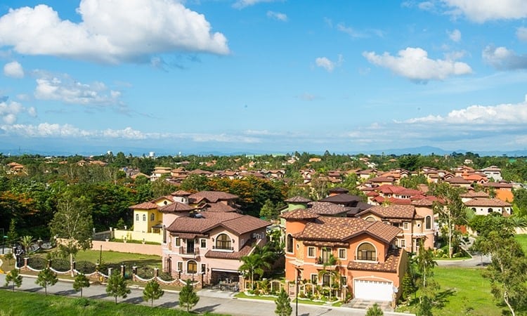 An spacious sprawling wide area of an italian inspired subdivision with clean beautiful houses and lots in warm welcoming colors, classic accents, pine trees, lush greenery, healthy grass, and gates of rustic bricks