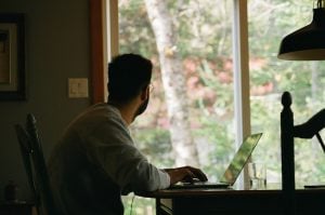 A back-view photo of a man wearing a gray sweater; the backdrop consists of an outdoor view of an indoor home office. 