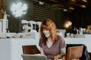 Photo of a smiling woman sitting on a brown wooden chair while using her laptop - Real estate agents - Luxury Homes by Brittany