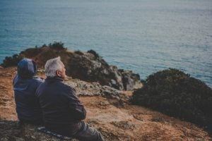 Two persons sitting on a rock staring at the body of water during daytime celebrating grandparents month | Luxury Homes by Brittany Corporation