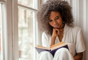 black woman taking a break by reading a book by a window | Luxury homes by brittany corporation