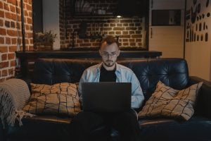 man using his silver laptop while sitting on a black leather couch | Why Real Estate is the Best Investment in 2022