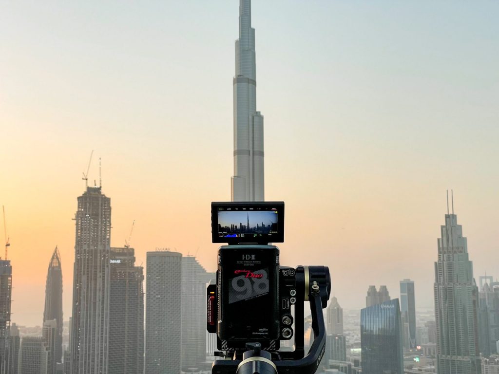 Black camera on a tripod. On the backdrop are tall buildings under daylight